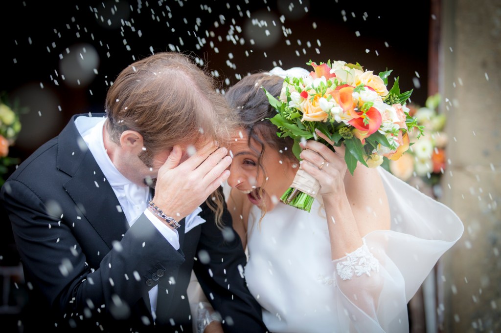 Bride and groom laughing and shielding themselves from confetti at their wedding ceremony.