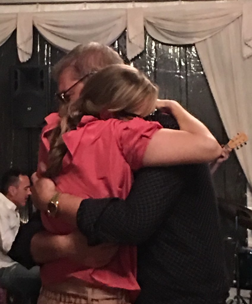 A heartfelt moment of a couple embracing at a wedding celebration, surrounded by guests and musical instruments in the background.
