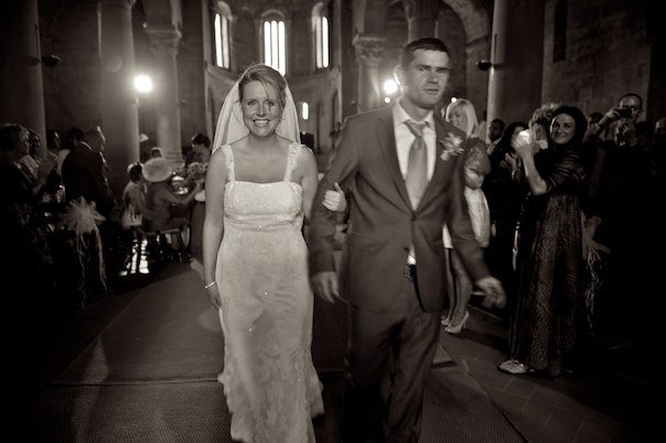 Bride and groom walking down the aisle in a historical church in Tuscany, with guests cheering in the background.