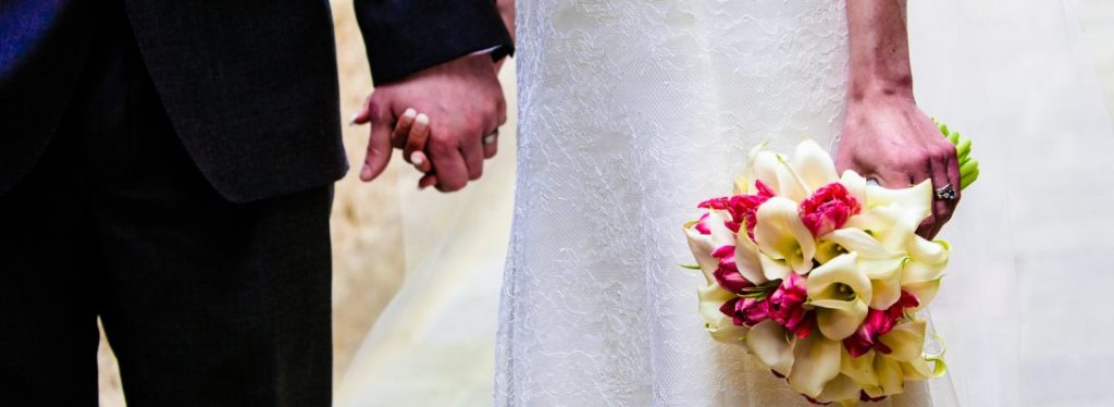 Close-up of a couple's hands holding each other, showcasing their wedding rings and the bride's bouquet of white and pink flowers.
