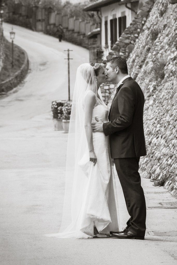 A bride and groom share a kiss on a charming street in Tuscany, surrounded by rustic buildings and a gently sloping road.