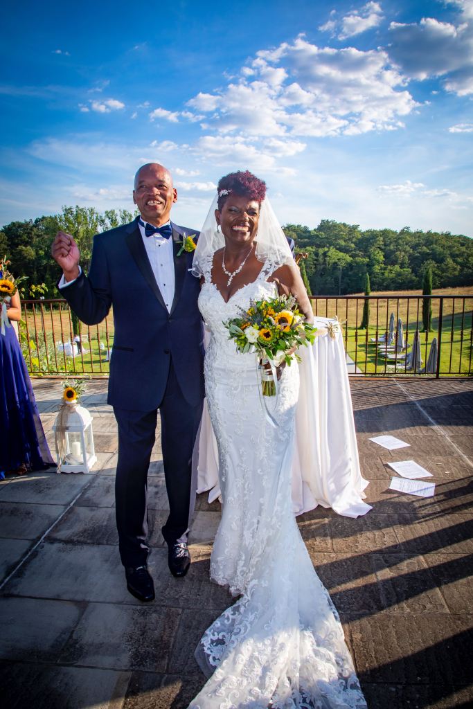 A joyful couple poses together outdoors in Tuscany, surrounded by a scenic backdrop featuring trees and a clear blue sky, with the bride holding a bouquet of sunflowers.