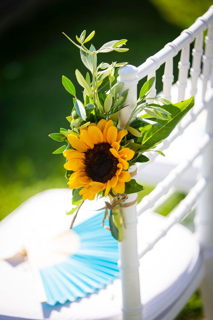 A decorative wedding chair adorned with a vibrant sunflower and greenery, alongside a blue hand fan in the background.